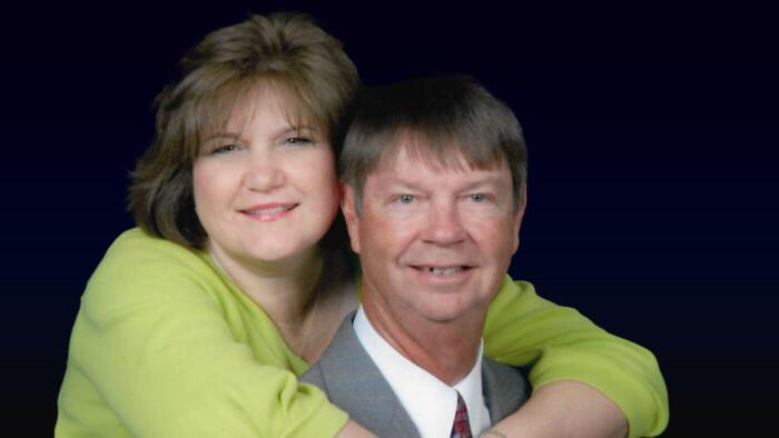 A head-and-shoulders portrait against a dark background of a man in a gray suit with a woman in a green top embracing him from behind with her arms and hands crossed over his shoulders.